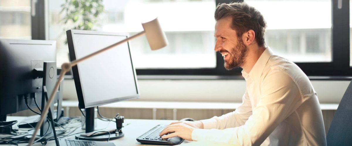 A man smiling while working at an office desk with a computer and natural daylight streaming in through large windows.